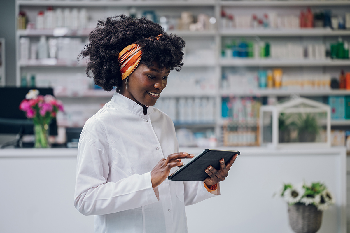A young multiracial professional healthcare service worker is st A cheerful young African-American doctor is standing in a drugstore with a tablet in her hands and scrolling while smiling at it. A multiracial drugstore worker is doing studies on medicament.
