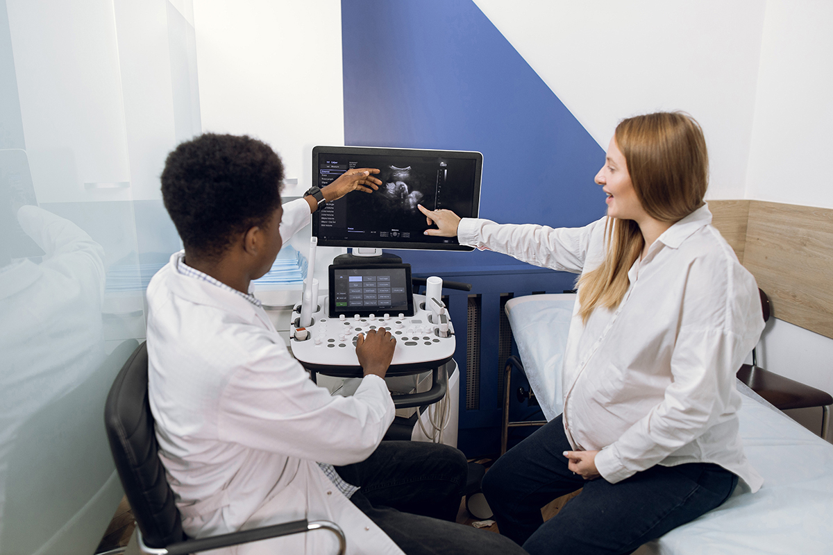 Beautiful young pregnant woman in modern clinic, pointing on the screen of ultrasound machine and asking her male African doctor obstetrician about the results of ultrasound test. Beautiful young pregnant woman in modern clinic, pointing on the screen of ultrasound machine and asking her male African doctor obstetrician about the results of ultrasound test..