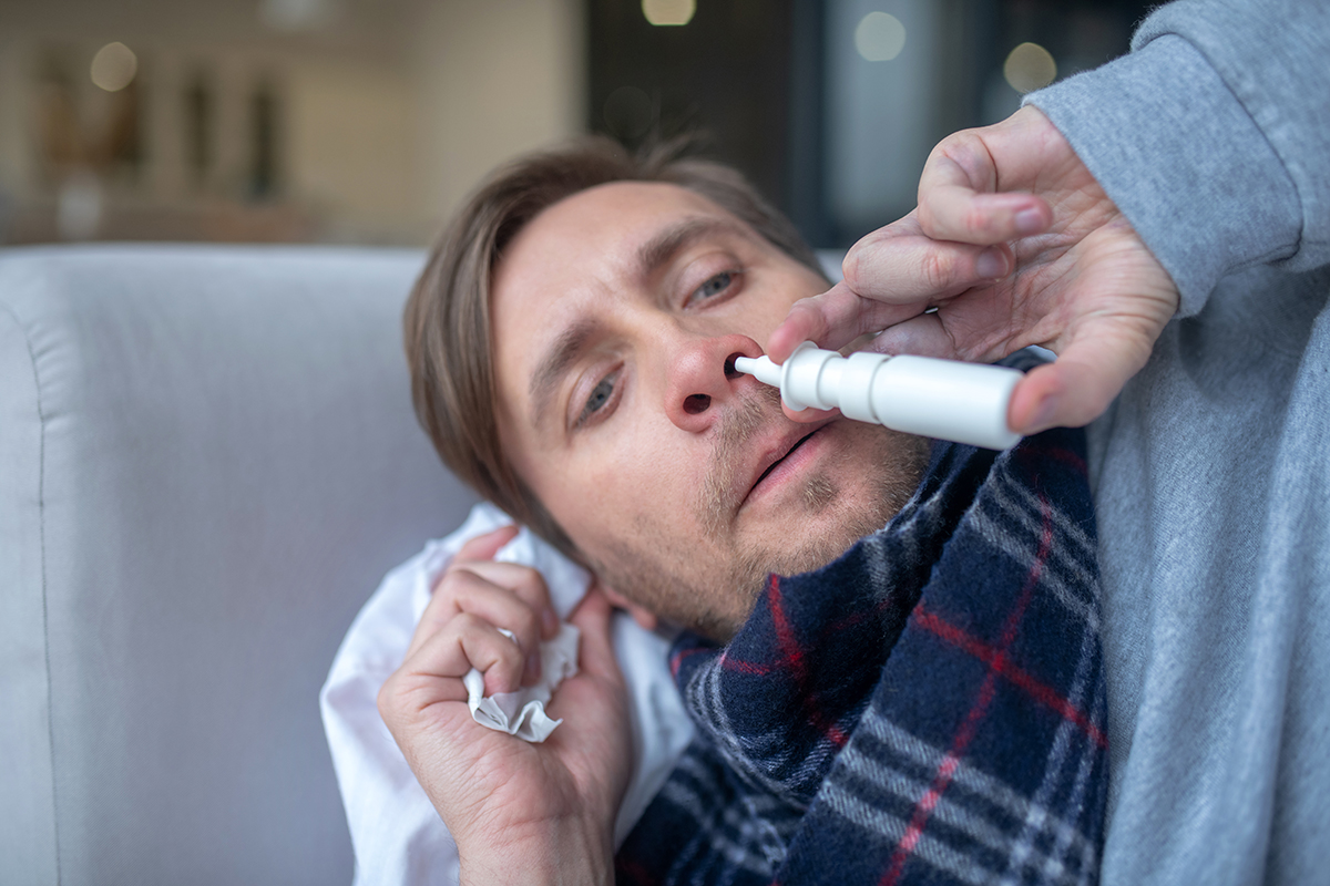 Blue-eyed man having stuffy nose taking nasal spray Having stuffy nose. Blue-eyed man having stuffy nose and headache taking nasal spray