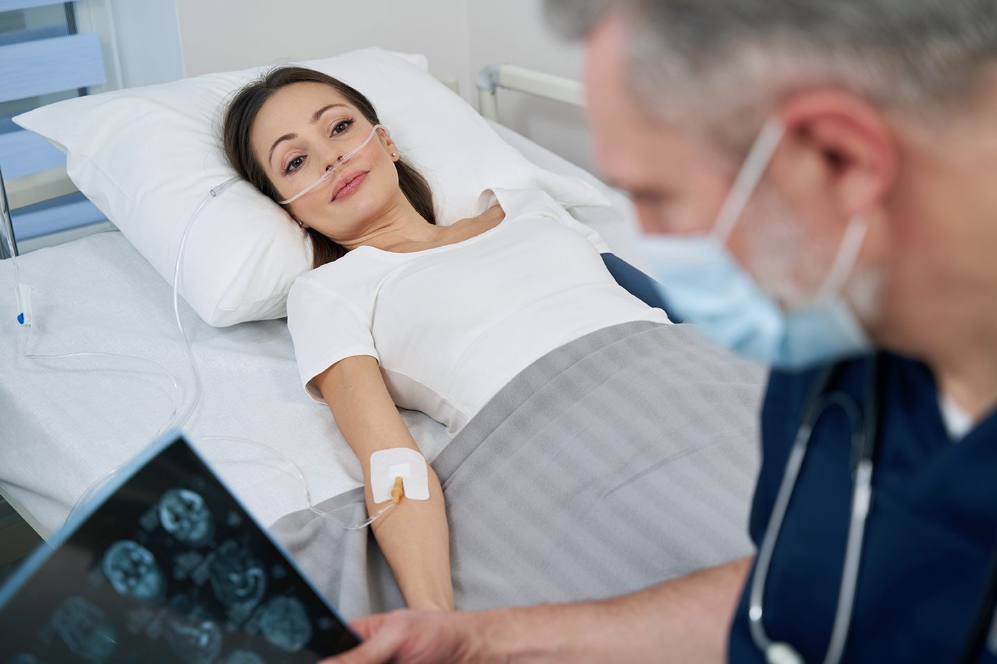Focused neurologist examining MRI scans of young woman Male physician seated on hospital bed beside female patient looking at her brain images