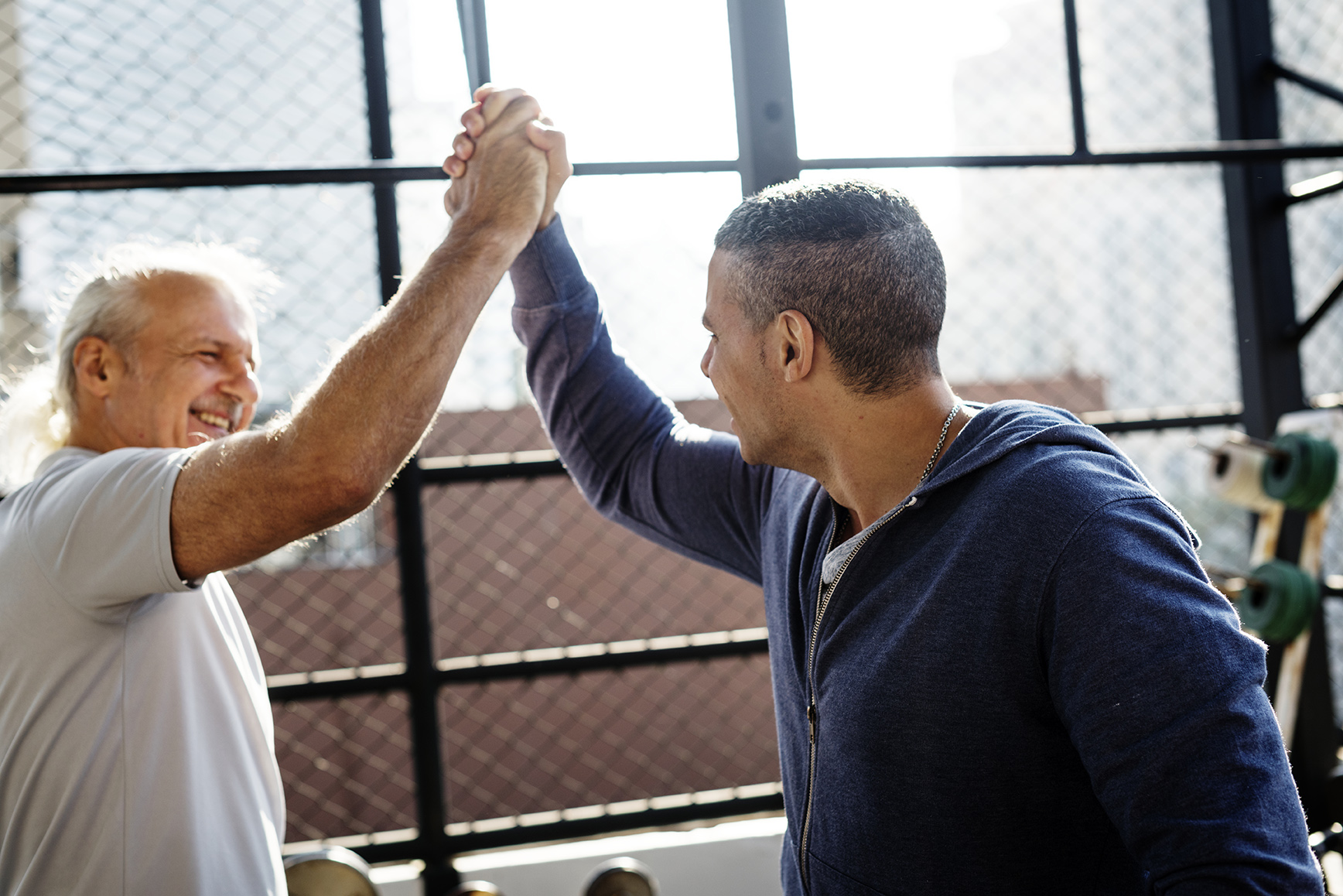People exercising at fitness gym Two men at the gym grabbing hands and celebrating. There is a window in the back and a chain fence.