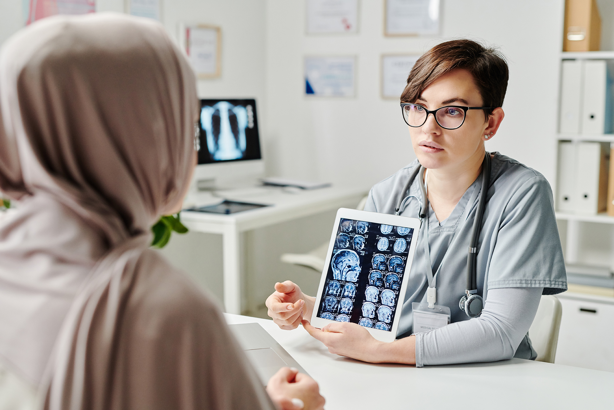 Young confident radiologist in uniform commenting brain scan to patient Young confident radiologist in uniform commenting brain scan on tablet screen to Muslim female patient in hijab sitting in front of her