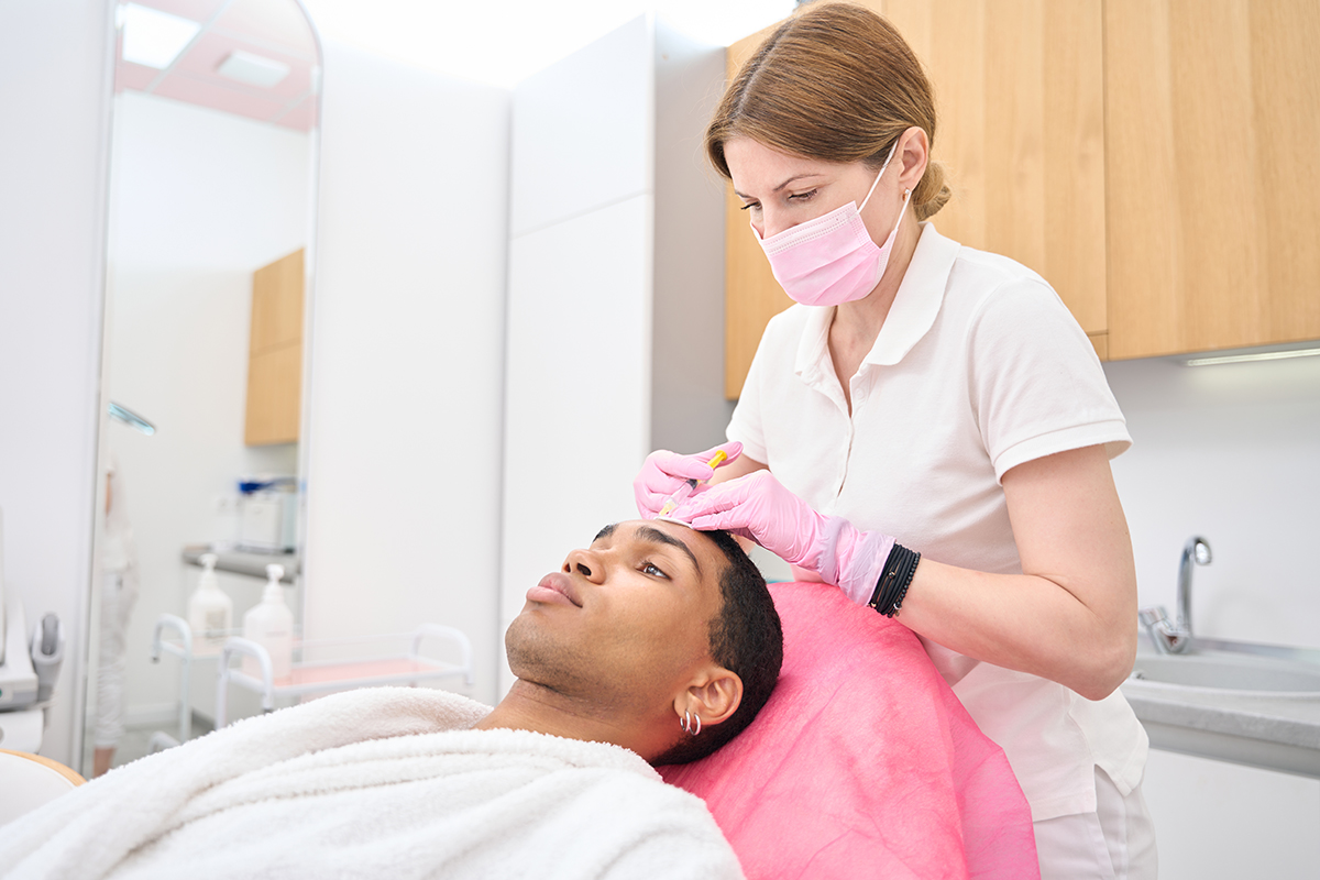 Beauty salon cosmetician performing mesotherapy procedure on customer Esthetician in nitrile gloves and face mask administering dermal filler injection into young man forehead