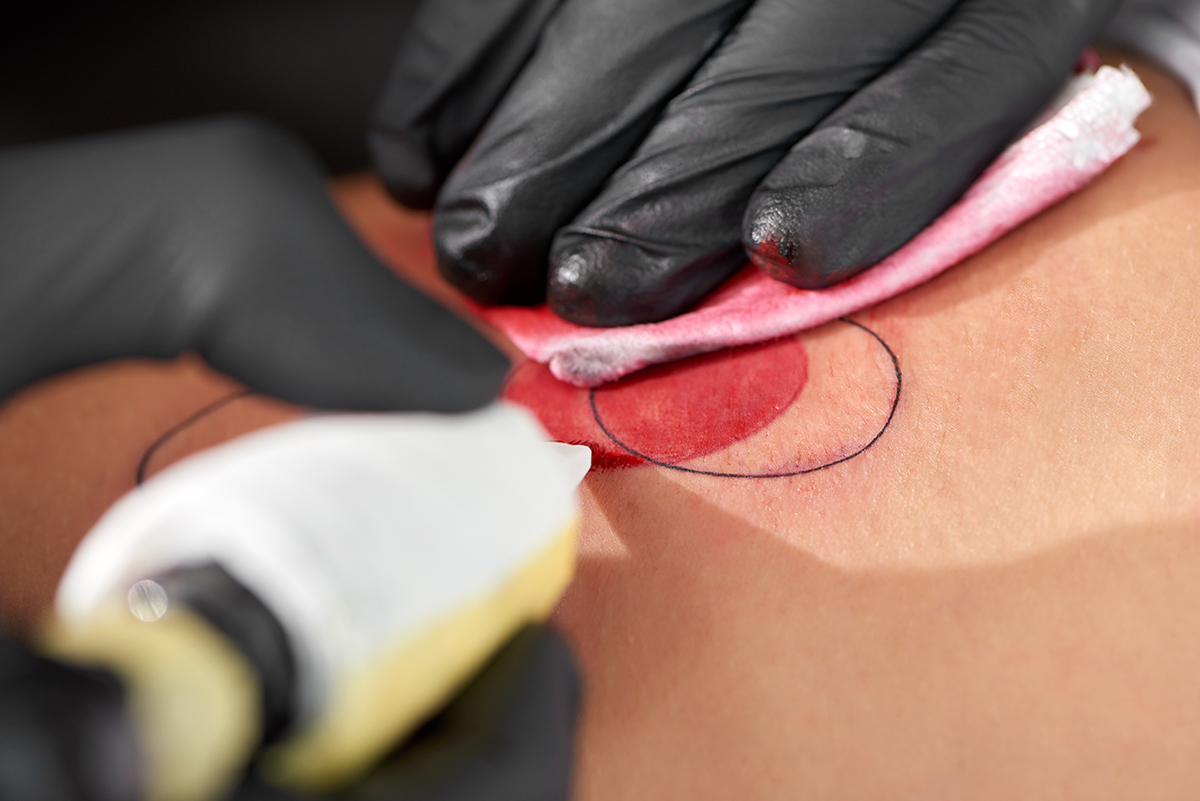 Close up of isolated male hands with tattoo machine during working. Master in black gloves doing pattern on naked skin and using black and red ink. Concept of advertising tattoo salon and its worker.