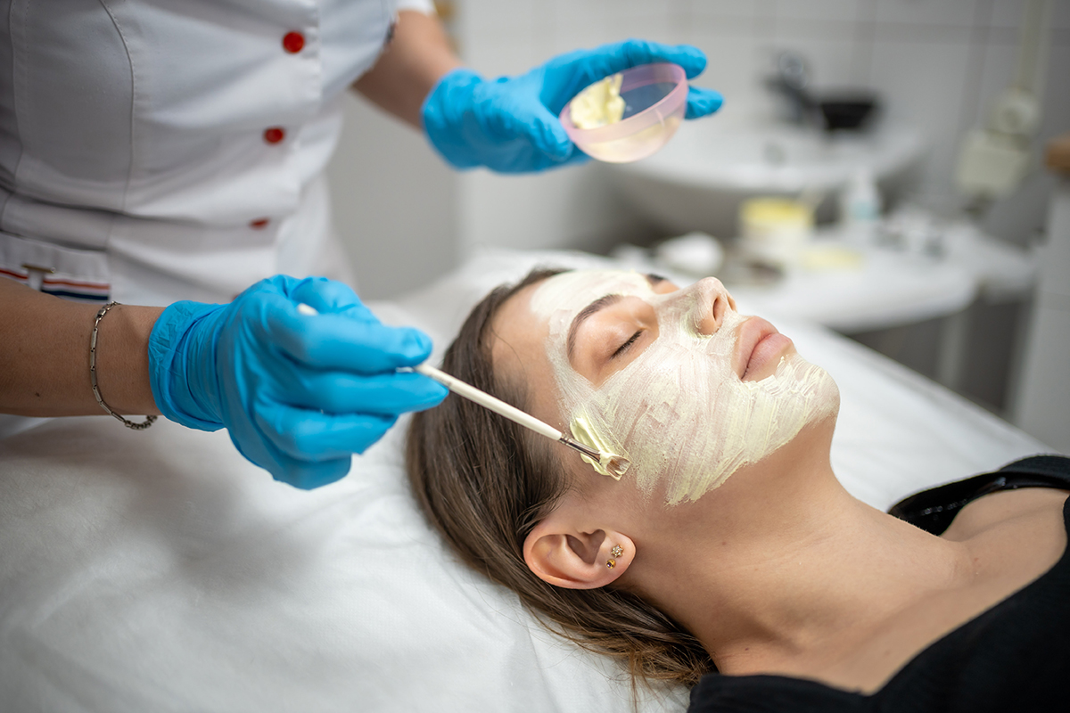 Doctor cosmetologist or dermatologist making face mask in cosmetology salon. Professional Beautician applying face mask on caucasian woman face lying on bed in bathrobe. Doctor cosmetologist or dermatologist making face mask in cosmetology salon. Professional Beautician applying face mask on caucasian woman face lying on bed in bathrobe. High quality photo