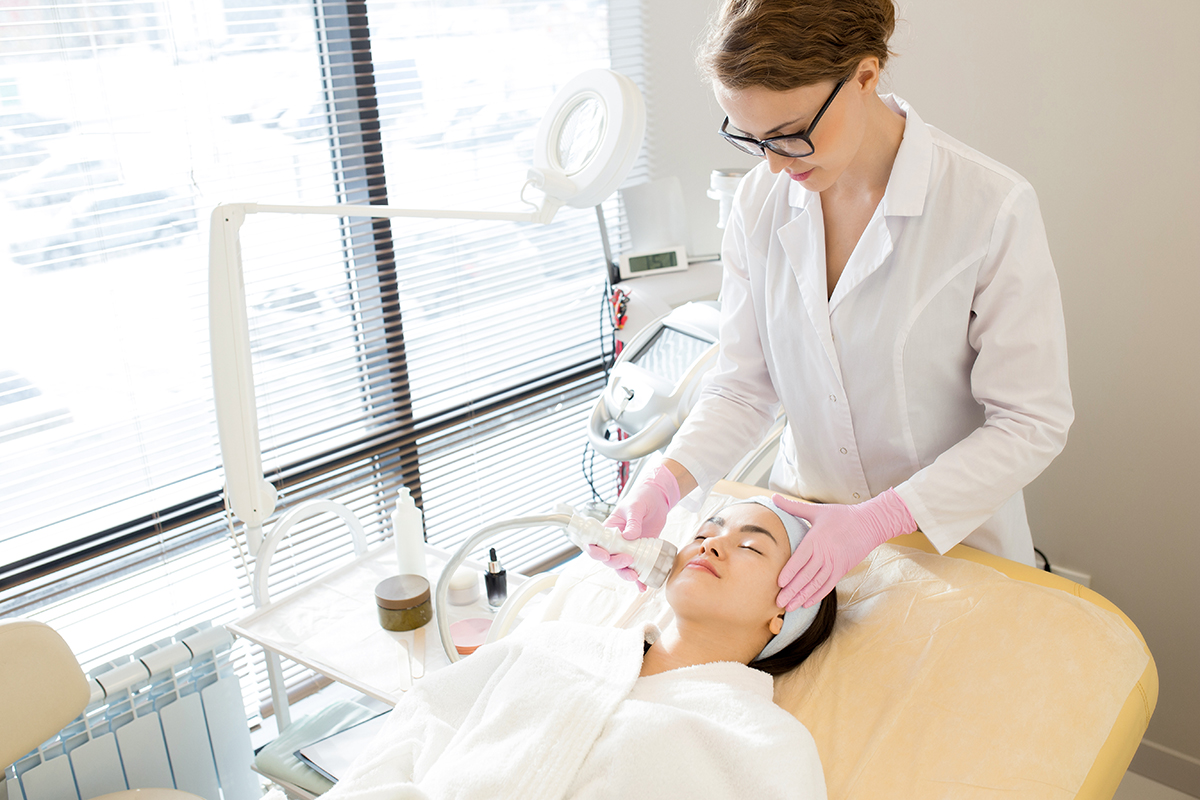 Enjoying Facial Massage Relaxed young woman wearing bathrobe lying on treatment table while highly professional beautician massaging her face with special equipment, interior of modern facial room on background