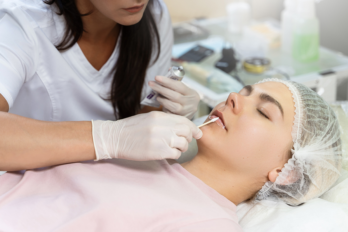 Professional permanent makeup artist applying anesthetic on client's lips before lip blushing procedure