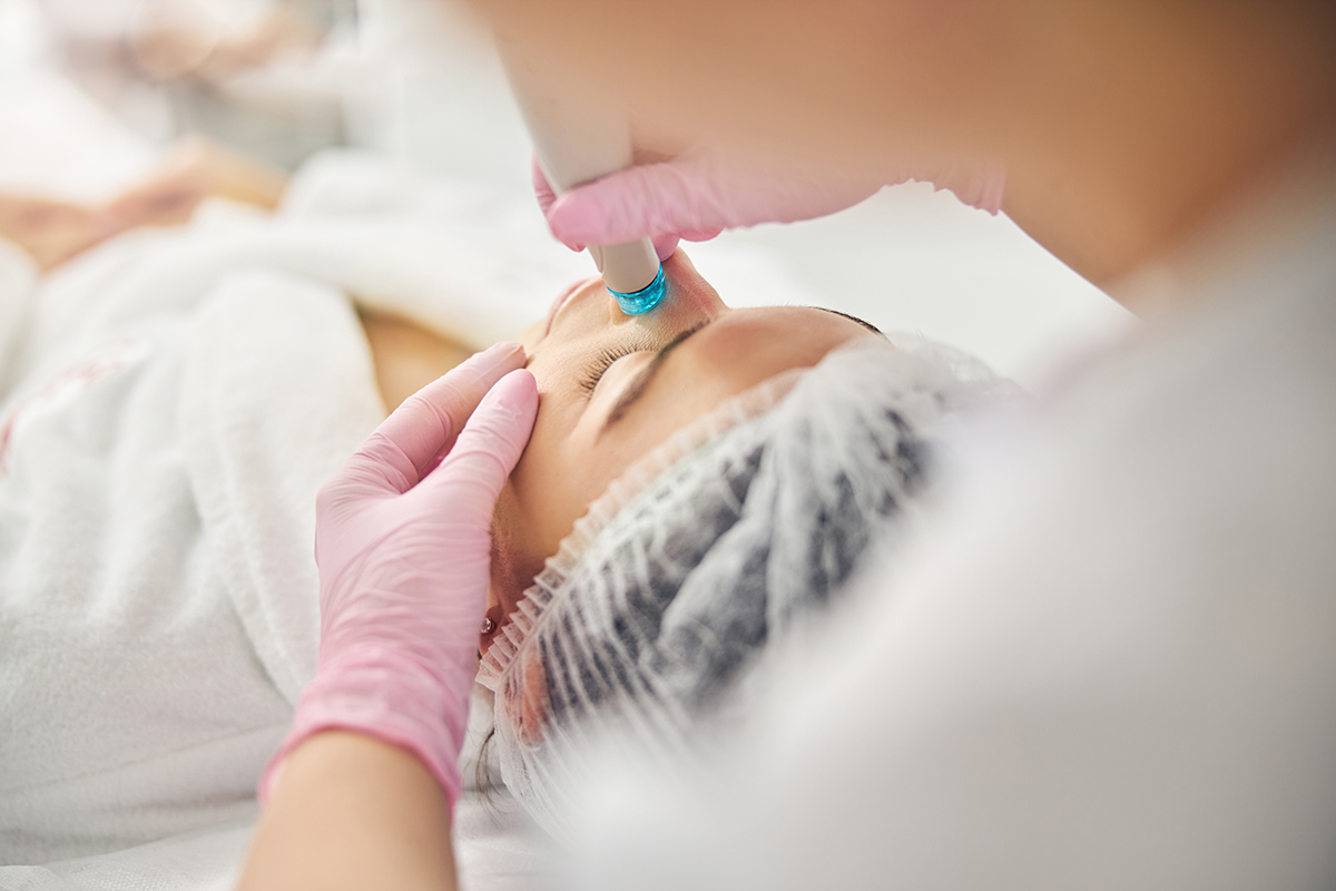 Young Caucasian female undergoing a dermatological procedure Dark-haired woman in a disposable gauze cap being treated by a doctor in latex gloves