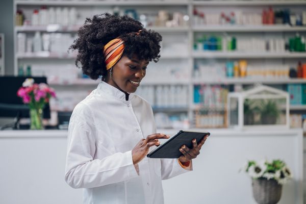 A cheerful young African-American doctor is standing in a drugstore with a tablet in her hands and scrolling while smiling at it. A multiracial drugstore worker is doing studies on medicament.