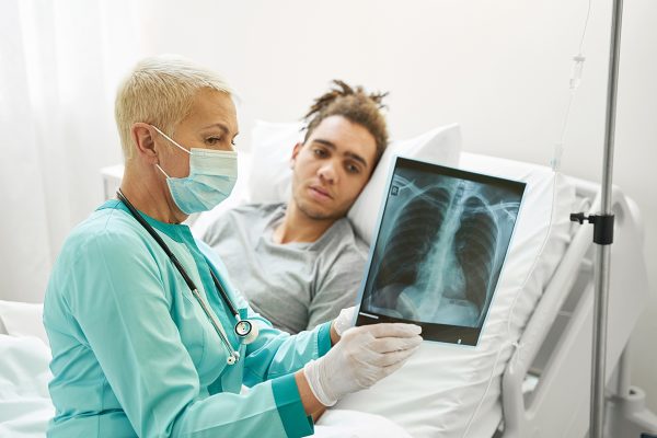Senior medical person demonstrating chest radiograph to male patient, lying in bed of health care center