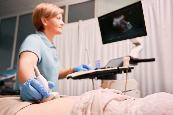 Focus on female physician's hand in sterile glove examining woman with ultrasound scanner. Doctor moving ultrasound transducer on patient abdomen, looking at display. Concept of medical examination.
