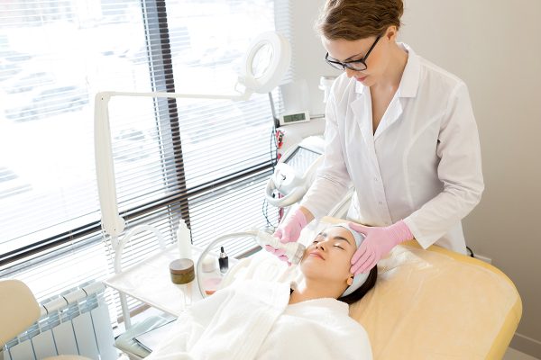Relaxed young woman wearing bathrobe lying on treatment table while highly professional beautician massaging her face with special equipment, interior of modern facial room on background