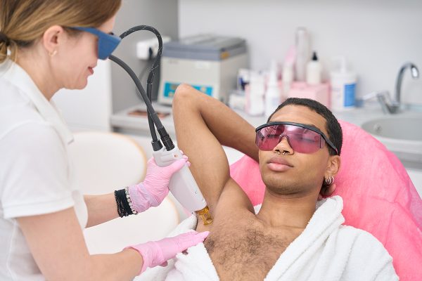 Beautician in nitrile gloves and safety goggles removing unwanted hair from man armpit using modern equipment