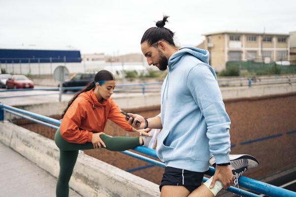 Active man wearing sports clothes using his phone and stretching outdoors after working out. His female friend is stretching too.