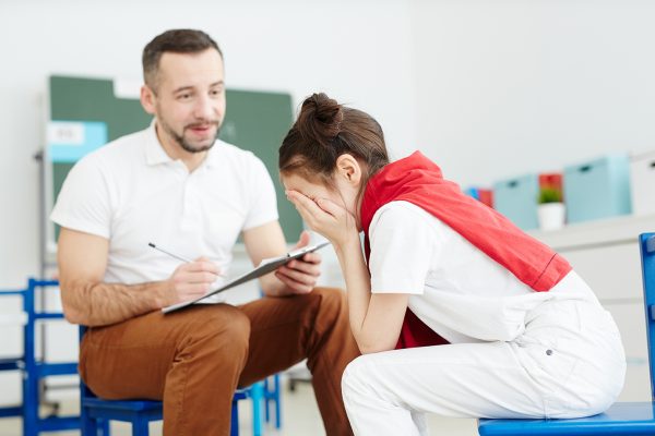 Crying girl sitting on chair in front of school psychologist or teacher trying to comfort her