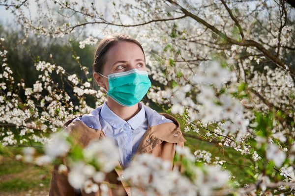 Woman allergic suffering from seasonal allergy at spring, posing in blossoming garden at springtime, wearing medical mask among blooming trees. Spring allergy concept