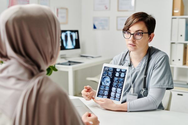 Young confident radiologist in uniform commenting brain scan on tablet screen to Muslim female patient in hijab sitting in front of her