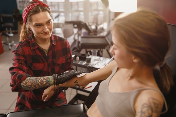 Woman tattoo artist wering medical gloves sitting next to client and applying gel on her hand with napkin